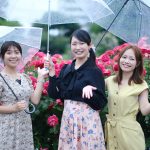 雨のなか女性３人がバラがたくさん咲いてる前で傘をさしポーズ
